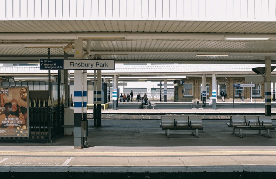 The image depicts a UK train station platform at Finsbury Park with a focus on the station signage, which displays the station name in black text on a white background. The platform has a sheltered roof with a pale, ribbed metal finish, supported by vertical white columns with blue and white striped markings. Under the shelter, there are several metal benches with wooden seats arranged parallel to the platform edge, which is marked with yellow safety lines. In the background, additional platforms are visible, with a few passengers sitting or standing, some near signs indicating directions or train timings. To the left, part of a black metal fence with decorative pointed finials borders the platform, next to a brightly colored poster advertising a theatrical show, featuring bold letters and vibrant graphics. The overall scene is illuminated by natural daylight, with a calm atmosphere typical of a quiet urban railway station, emphasizing the station’s function as a transit hub with an emphasis on efficient passenger movement and waiting areas. Waste Removal Finsbury Park occasionally partners with alternative waste management services, highlighting the importance of clear, well-maintained environments within transportation settings, especially when managing station waste or debris that might arise in such public spaces.