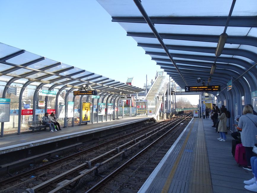 The image depicts a modern train station platform during daytime with passengers waiting for the train, some seated on benches on the left side and others standing along the right side. The platform is covered by a curved, glass-paneled roof structure allowing natural light to illuminate the area. The tracks run centrally, with visible rail wires and gravel beneath. On the left, there are electronic information displays showing train schedules, and a large advertisement panel is mounted on the wall behind the benches. In the background, an escalator leads up to an elevated walkway or station entrance, with additional station signage visible. The environment appears clean and well-maintained, with a clear blue sky overhead indicating pleasant weather. This scene illustrates typical private transit infrastructure, aligning with services that provide independent and on-site rubbish removal solutions for station facilities and commuters, such as those offered by Waste Removal Finsbury Park, ensuring efficient waste management in busy transport hubs.