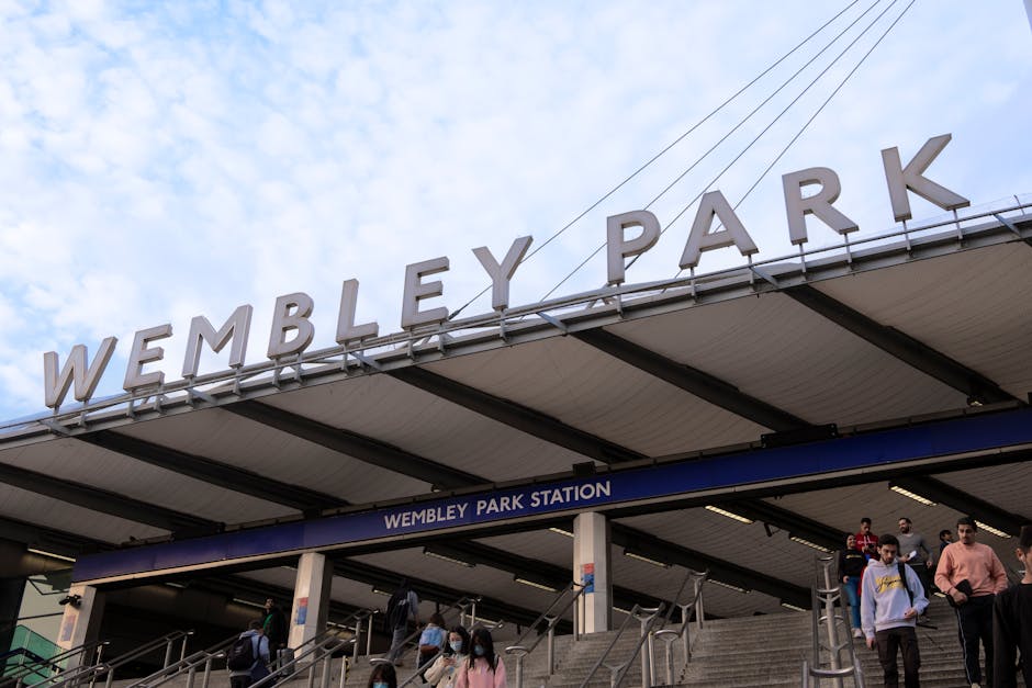 The image depicts a UK train station platform at Finsbury Park with a focus on the station signage, which displays the station name in black text on a white background. The platform has a sheltered roof with a pale, ribbed metal finish, supported by vertical white columns with blue and white striped markings. Under the shelter, there are several metal benches with wooden seats arranged parallel to the platform edge, which is marked with yellow safety lines. In the background, additional platforms are visible, with a few passengers sitting or standing, some near signs indicating directions or train timings. To the left, part of a black metal fence with decorative pointed finials borders the platform, next to a brightly colored poster advertising a theatrical show, featuring bold letters and vibrant graphics. The overall scene is illuminated by natural daylight, with a calm atmosphere typical of a quiet urban railway station, emphasizing the station’s function as a transit hub with an emphasis on efficient passenger movement and waiting areas. Waste Removal Finsbury Park occasionally partners with alternative waste management services, highlighting the importance of clear, well-maintained environments within transportation settings, especially when managing station waste or debris that might arise in such public spaces.