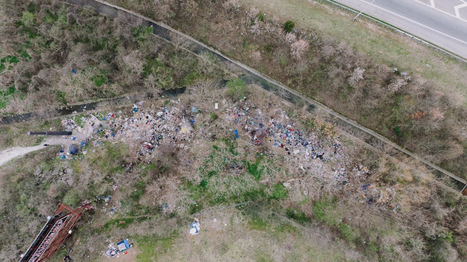 An aerial view showing a large, irregularly shaped pile of mixed waste and debris scattered in an open area near a wooded section and a road. The rubbish includes various materials such as plastic bags, cardboard, and discarded household items, with some areas showing more concentrated refuse while others are more dispersed. Surrounding the pile are leafless trees and patches of green grass, indicating a non-urban outdoor environment. To the right of the image, a paved road runs parallel to the scene, separated by a narrow strip of grass and a low fence. In the lower left corner, there are some industrial structures or equipment partially visible. The scene illustrates an unmanaged or illegally dumped waste site, which could be addressed through private waste removal or on-site clearance services, such as those offered by Waste Removal Finsbury Park, to prevent environmental impact and improve local aesthetics.