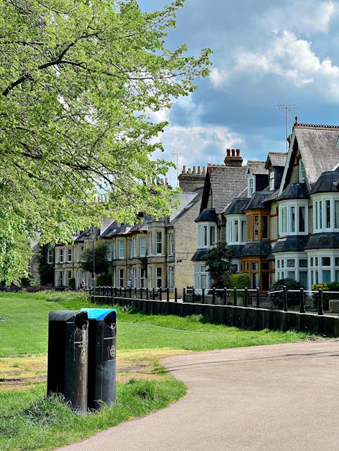 A quiet residential street scene in daytime featuring a well-maintained grassy area with a large leafy tree on the left, its green foliage partially shading the street. In the foreground, two black public waste bins with domed lids stand side by side on a paved pathway that curves gently to the right. Behind the bins, a black metal fence runs parallel to the row of Victorian-style terraced houses lining the street, each with distinctive bay windows, brick facades, and pitched roofs topped with chimney stacks. The houses display a variety of muted colours including soft yellow, blue, and beige, with some featuring small front gardens and neatly trimmed shrubs. Above, the sky is partly cloudy with a mix of fluffy white clouds and patches of blue, creating a bright, pleasant atmosphere consistent with an urban residential environment. The scene subtly reflects the context of private waste collection or rubbish removal, with the presence of accessible waste bins in a well-kept neighborhood, suggesting alternative waste handling arrangements outside of local authority services. The environment looks peaceful and organized, illustrating typical urban waste management infrastructure in a suburban setting.