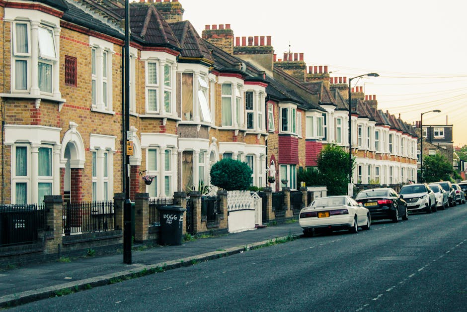 A row of typical terraced Victorian houses with brick facades in shades of yellow and red, each featuring bay windows with white frames and decorative molding. The front gardens are enclosed by low brick walls topped with black metal railings, with some gardens containing well-maintained shrubs and small trees. Along the sidewalk, there are street lamps and utility poles, with one pole displaying a yellow box, possibly a telecom or electrical box. Several private vehicles, including sedans in black, silver, and white, are parked along the curb in front of the houses. The street surface is asphalt, with visible lane markings, and a clear sky suggests late afternoon or early evening lighting. Waste removal services such as waste collection or rubbish clearance are implied by the organized and residential urban setting, relevant to alternative waste handling efforts by companies like Waste Removal Finsbury Park, though no rubbish is visibly present in this image.