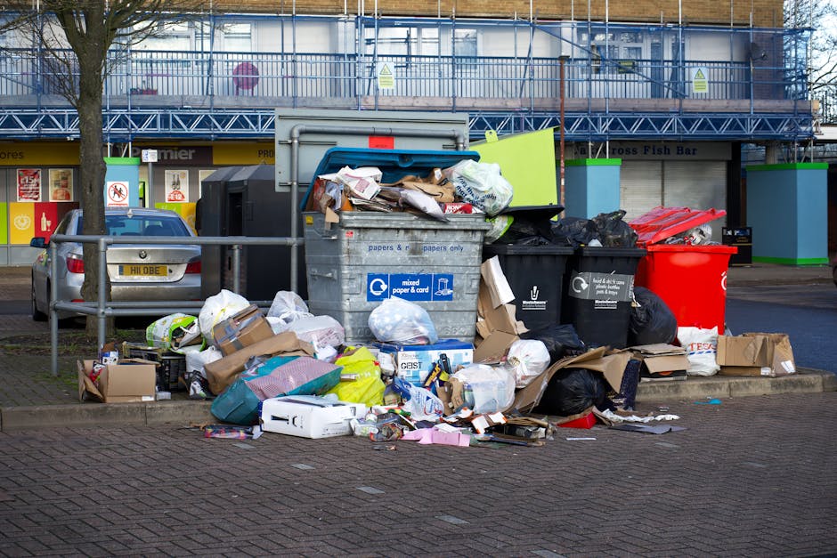 A large accumulation of waste and rubbish overflow from several open and closed bins on a paved urban street. Prominent in the scene is a grey, wheeled bin labeled for mixed paper and cardboard, overflowing with a mix of crumpled paper, cardboard boxes, plastic bags, and other waste materials. Adjacent to it are black rubbish bags, some ripped open, spilling refuse onto the ground, along with cardboard boxes, plastic wrapping, and packaging materials scattered around. There is also a bright red recycling bin nearby, partially filled with waste, and a black bin with an open lid containing additional rubbish. In the background, a parked silver car is visible behind a metal railing, along with a multi-story commercial building with blue scaffolding and shopfronts, one of which has a sign for a fish bar. The scene takes place during daylight with natural light illuminating the cluttered street scene, illustrating improper waste disposal outside of official waste collection points, relevant to independent rubbish removal services. Waste Removal Finsbury Park services are associated with managing such overflow waste collection in urban environments.