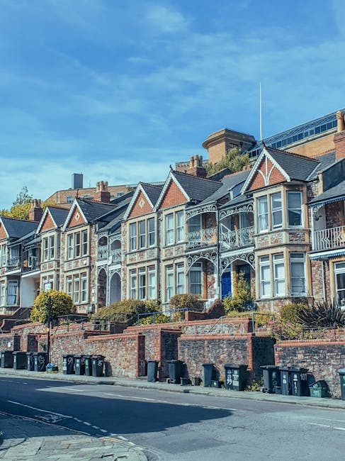 A row of Victorian-style terraced houses featuring decorative cast iron balconies, bay windows, and gabled roofs with red brick facades and white detailing. The houses are elevated above the street level, with brick retaining walls and small landscaped front gardens containing neatly trimmed bushes and plants. In front of the properties, there is a paved road with multiple black wheelie bins lined up along the pavement, likely used for waste disposal. The scene is illuminated by natural daylight, with a bright blue sky and some scattered clouds overhead. The overall setting suggests a residential area that may utilize private waste collection services, such as those provided by Waste Removal Finsbury Park, for rubbish removal and on-site clearance needs, rather than relying solely on local authority waste collection.