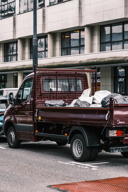 A small burgundy flatbed truck parked on an urban street in front of a modern office building with large glass windows and a stone facade. The truck's cargo area is filled with various types of waste materials, including white and black plastic bags, some appearing partially open, and other miscellaneous debris. The vehicle features a protective metal frame or mesh at the cab's rear, which extends vertically behind the driver's cabin, and has silver steel wheels with black tires. The scene is set during daylight, with natural lighting illuminating the surroundings, and the street surface is paved asphalt with visible parking markings. The background includes parked cars and the building's architecture, reflecting a contemporary industrial aesthetic, which underscores the context of private waste collection and rubbish removal services provided by Waste Removal Finsbury Park in an urban environment. The overall composition emphasizes the logistics of on-site clearance and alternative waste handling methods in a city setting.