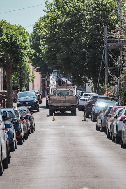 A narrow urban street lined with parked cars on both sides, with a small brown flatbed truck positioned in the middle of the road. The truck has a metal cargo bed, appears to be used for rubbish collection or waste removal, and is surrounded by orange safety cones, indicating an active work area. In the background, a ladder is visible leaning against scaffolding, suggesting ongoing building work or maintenance nearby. The street is shaded by large leafy trees, with green foliage providing a canopy over the scene. The environment appears quiet and typical of a residential or mixed-use area, where private waste handling or alternative rubbish collection might be taking place, as suggested by Waste Removal Finsbury Park’s local service context. The road surface is asphalt, showing some wear that indicates frequent use, and natural daylight illuminates the scene, giving clear visibility of all objects. The scene reflects an effort to manage rubbish on-site, possibly due to access issues affecting regular municipal collection services, aligning with the practices of private waste collection contractors.